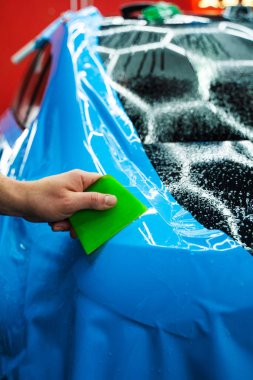 Mavi PPF, yeşil cam sileceği ile arabayı temizlerken ve hasarı engellemek için uygulanmış. Yeşil bir cam sileceği kullanarak yüksek parlak mavi bir PPF uygulamak, arabanın baloncuksuz ve pürüzsüz bir yüzey sağlamasını sağlar