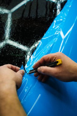 Mavi PPF 'yi bıçakla kesiyorum. Detaylı iş sırasında mavi boya koruma filmini bıçakla kesmek hassas kenarlar sağlar. Detaylı teknisyen mükemmel kontur için bir araba paneli üzerindeki mavi PPF buduyor