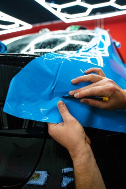 Mavi PPF 'yi bıçakla kesiyorum. Detaylı iş sırasında mavi boya koruma filmini bıçakla kesmek hassas kenarlar sağlar. Detaylı teknisyen mükemmel kontur için bir araba paneli üzerindeki mavi PPF buduyor