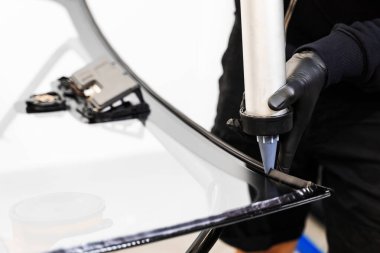Bir arabanın ön camına kabartma tabancası kullanarak yapıştırıcı sürüyorum. Urethane kazıma silahı, arabanın ön camının değiştirilmesinde su geçirmez bir mühür oluşturarak esnek bir yapıştırıcı uyguluyor.