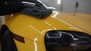 Tamirci, arabanın 4K görüntüsünü silmek için gri bir mikrofiber bez kullanıyor. Temizlikçi, sarı bir arabanın vücudunu mikrofiber bezle siler..