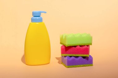 Stack of multi-colored dish wash sponges with bottle of soap on pastel orange background. Household cleaning scrub pad. Home cleaning concept