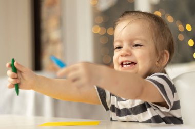 Happy cute caucasian baby girl sitting at the table at home. Smile and looking at hands with crayons.