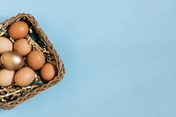 Gift basket with easter eggs, golden and natural color eggs in basket. Top view on eggs above pastel blue background