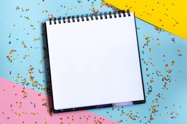 Pastel mavi arka plan ve renkli konfeti üzerine beyaz kağıt içeren boş spiral çizim defterinin taklidini yap. Temiz sayfalı açık defterin üst görünümü. İleti için şablon. Boşluğu kopyala