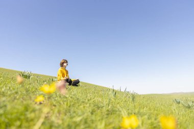 Genç bir insan yeşil bir yamaçta sakince oturuyor. Etrafı kır çiçekleriyle çevrili. Sakin ve rüya gibi bahar atmosferi. Barış ve doğa ile bağlantı.