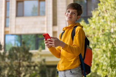Sarı kapüşonlu genç bir çocuk akıllı bir telefon tutuyor ve doğrudan kameraya bakıyor. Tarafsız arka plan, gelişigüzel poz ve modern teknoloji. 