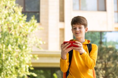 Genç bir çocuk yüksek katlı bir apartmanın önünde duruyor, elinde akıllı bir telefon var ve ekrana bakarken gülümsüyor. Modern dijital hayatı, iletişimi ve şehir yaşam tarzını temsil eder. 