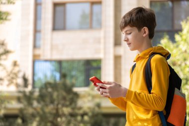 Genç bir çocuk elinde akıllı bir telefonla bir apartmanın önünde durup gülümsüyor. Dijital erişilebilirliği, çevrimiçi iletişimi ve modern bağlı yaşam tarzını sembolize eder.