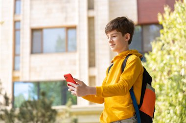 Ergen çocuk akıllı bir telefon tutuyor ve yüksek bir binanın önünde gülümsüyor. Teknolojinin iletişim, öğrenme ve modern hayatı şekillendirdiği dijital çağı sembolize eder.. 