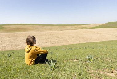 Açık arazide yeşil çimlerin üzerinde oturan ve mesafeye bakan birinin yan görüntüsü. Metin veya tasarım için kopya alanı olan barışçıl açık hava sahnesi.