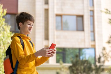Genç çocuk yüksek bir binanın yanında duruyor, akıllı telefonuna bakıyor ve gülümsüyor. Şehirdeki kentsel yaşam tarzı, dijital bağlantı ve günlük yaşamı temsil eder.. 
