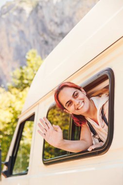Kamp arabasının camından el sallayan genç bir kadın. Minibüs tatili ve açık hava yaz macerası. Göçebe yaşam tarzı konsepti