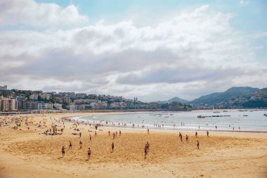 Donostia San Sebastian, İspanya 'da güneşli bir günde kumlu La Concha plajında apartman binalarına karşı yaz tatilinin tadını çıkaran bir kalabalık.