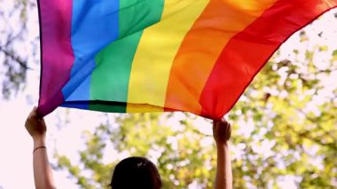 Arka planda ağaçların arasından süzülen güneş ışığıyla canlı bir gökkuşağı bayrağını kaldıran bir kadının yavaş çekim videosu. Bayrak LGBTQ + gurur ve kapsayıcılığı simgeliyor