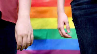 Lezbiyen bir çiftin arka planda canlı bir gökkuşağı gurur bayrağıyla el ele tutuşup LGBTQ + gurur ve kapsayıcılığı temsil ettiği yavaş çekim videosunu kapatın.