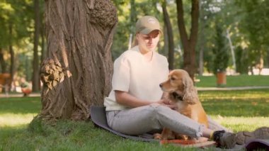 Genç bir kadın parktaki çimlerin yanında köpeğiyle oturur ve onu okşar. İngiliz Cocker Spaniel 'i ile Dışarıda Güneş Işığında Yürüyen Kız