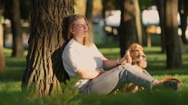Genç bir kadın parktaki çimlerin yanında köpeğiyle oturur ve onu okşar. İngiliz Cocker Spaniel 'i ile Dışarıda Güneş Işığında Yürüyen Kız