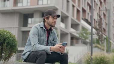 Şehir Binası 'nın Arkaplanına Karşı Otururken Günlük Giyimli Genç Adam Telefon Kullanıyor