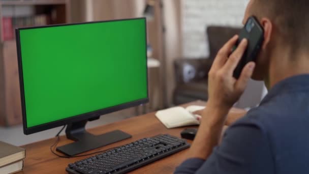 グリーンモックアップ画面でコンピュータで携帯電話の座席で話す若い男 Chromaキーディスプレイ付きデスクトップコンピュータ — ストック動画