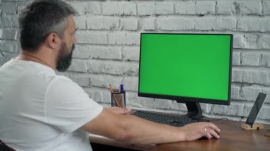 Sakallı Orta yaşlı adam, Bilgisayar Yeşil Ekran 'a bakar ve Defter' e bir şeyler yazar. Modern Ofiste Beyaz Tuğla Duvarlı Adam Çalışıyor