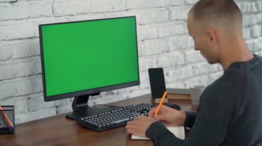 Adam Bilgisayar Yeşil Ekran 'a bakar ve Not Defterine Bir Şey Yazar. Genç Adam Beyaz Tuğla Duvarlı Modern Ofiste Çalışıyor
