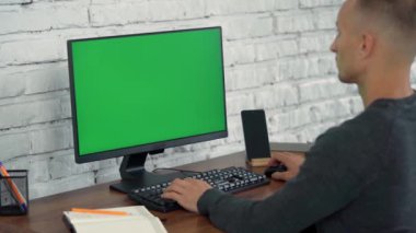 Adam Bilgisayar Yeşil Ekran 'a bakar ve Not Defterine Bir Şey Yazar. Genç Adam Beyaz Tuğla Duvarlı Modern Ofiste Çalışıyor