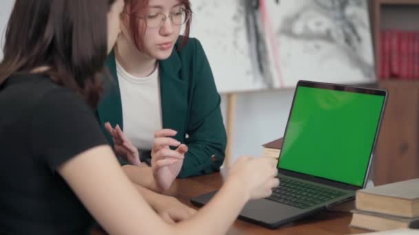 两个年轻的女孩互相交谈 看着笔记本电脑绿色屏幕 舒适室 图书馆的女学生交流 — 图库视频影像