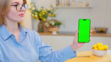 Gülümseyen Genç Kadın Yeşil Ekran Mock Up Ekranı ile Akıllı Telefon Gösteriyor. Arka plan, rahat ev mutfağı.