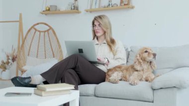 Gözlüklü genç kadın köpeğiyle koltukta otururken dizüstü bilgisayar kullanıyor. Arka plan... İç mekan.