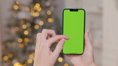 Yeşil Ekranlı Telefonun Yakın plan Çekimi Kadın Elleri Noel ağacının Arkasında ve Bulanık Işıklar. Kadın Kaydırma ve Dokunma Anahtarı Gösterimi Hareketleri Yapıyor