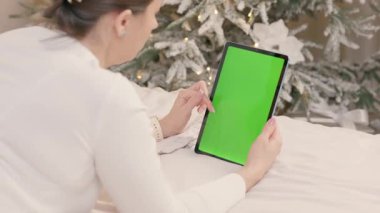 Bir kadın yeşil ekranlı tablet bilgisayar kullanıyor. Sıcak bir odada, Noel ağacı ve ışıklarıyla yatıyordu. İnternette Kız Sörfü ve Çevrimiçi Alışveriş