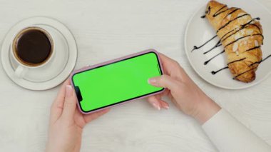 Kadın Ellerinde Yeşil Model Ekranlı Cep Telefonu 'nun Yakın Çekimi. Kafede dinlenirken akıllı telefon kullanan, kahve içen ve kruvasan yiyen bir kadın.