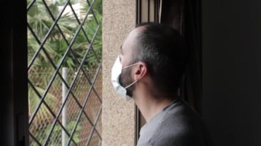 Man looks out the window with a mask to prevent getting coronavirus and throws the mask against the window, at home