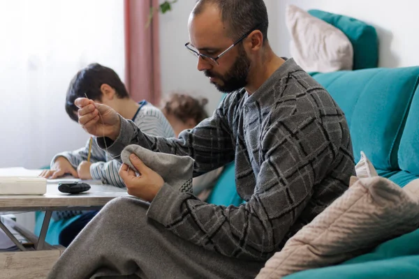 Man sewing a sock in the living room while his children do their homework