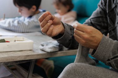 Man sewing a sock in the living room while his children do their homework