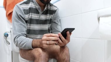 Pijamalı beyaz adam tuvalette oturuyor ve evdeki cep telefonuna bakıyor.