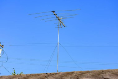 Uzun, eski moda bir çatı TV anteni, kablolarla desteklenen ve açık mavi gökyüzüne karşı kurulmuş fayanslı bir çatı üzerinde duruyor. Güç hatları