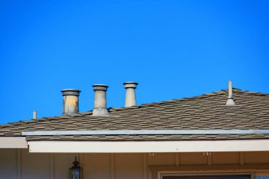 Açık mavi gökyüzünün altında kiremitli bir çatıdan birkaç çatı havalandırması yükseliyor. Havalandırmalar yıpranmış, pas ve kir birikmiş..