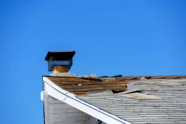 stock image A damaged roof with missing and peeling shingles, exposing wooden underlayment near a chimney topped with a metal chimney cap.