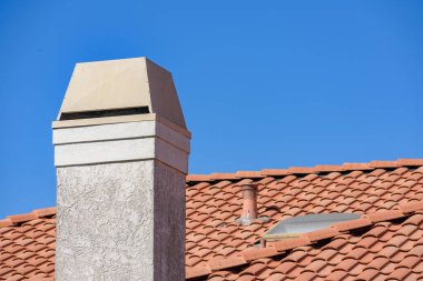 Kırmızı kiremitli çatıdan yükselen bej renkli metal kapaklı bir baca, parlak mavi bir gökyüzüne karşı kurulmuş. Arka planda küçük bir havalandırma borusu ve tavan penceresi görünür..