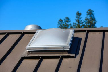 Kahverengi metal bir çatıda parlak, dikdörtgen bir tavan penceresi, açık mavi bir gökyüzü..