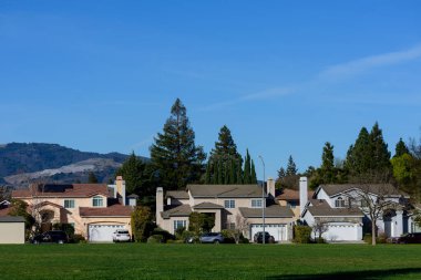 Modern evlerin, ön planda yeşil bir açık arazinin ve manzaralı dağ manzarasını çerçeveleyen uzun ağaçların bulunduğu yerleşim yerleri.