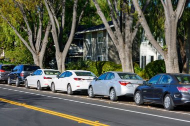 A row of parked cars along a residential street, shaded by tall trees, with houses and greenery in the background on a sunny day. Rear side view - California, USA - February 12, 2025 clipart