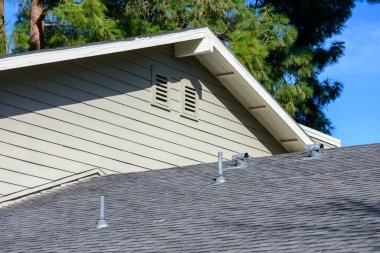 Eğri asfalt kiremit çatısında birden fazla metal menfez, bej kaplama ve tavan arası havalandırması var..