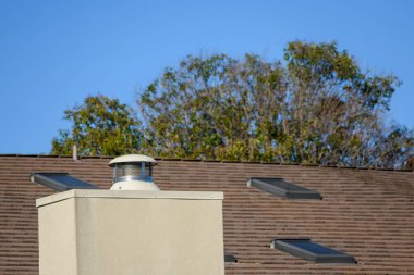 Bir yerleşim yerinin çatısı belirgin bir bej baca ve yapraklı ağaçlarla çevrili üç çatı penceresi içerir..