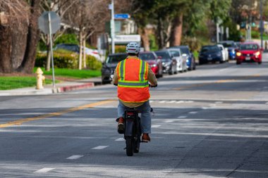 Parlak turuncu güvenlik yeleği ve kask giyen bir bisikletçi, park etmiş arabalar ve yemyeşil alanlarla çevrili, işaretli bir şehir caddesi boyunca elektrikli bisiklet sürüyor. Arka görünüm.