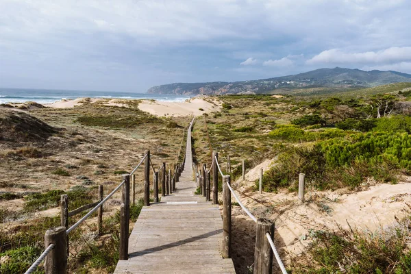 Empty, wooden boardwalk on a beach Praia do Guincho in Sintra. View of grass and sand with hills and Atlantic Ocean in the background