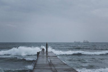 İki genç sörfçünün silueti fırtınalı bir günde dalgalanan dalgalarla rıhtımdaki beton platformda