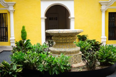 Sarı binanın yakınında yeşil bitkilerle çevrili ayrıntılı desenli bir çeşme.